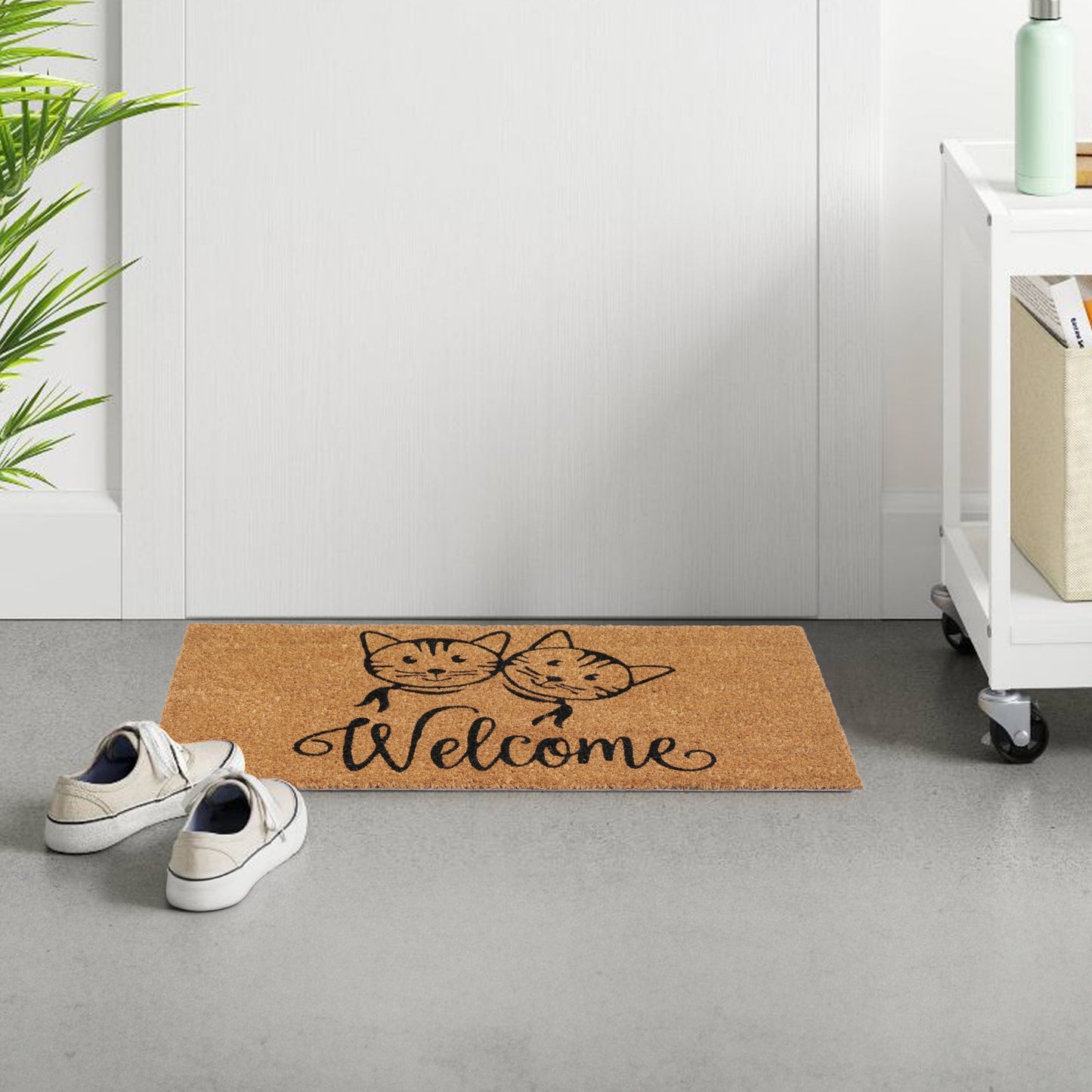 A rectangular natural coir doormat with a printed design featuring two cats and the word 'WELCOME' on a gray floor, next to a white door and a pair of shoes.