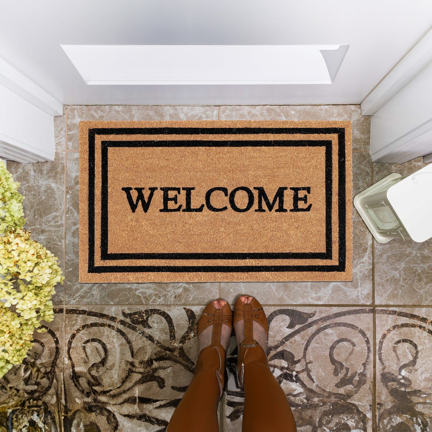 A coir doormat with 'Welcome' text and a decorative border pattern, placed on the floor by a doorstep.