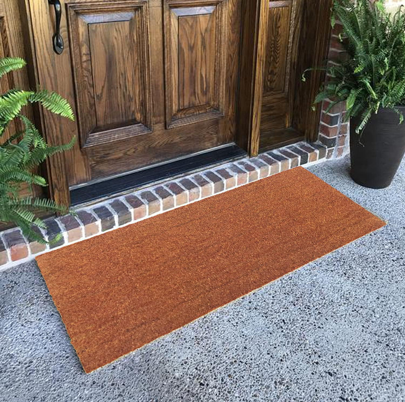 Brown plain coir doormat in front of a wooden door with plants on either side.