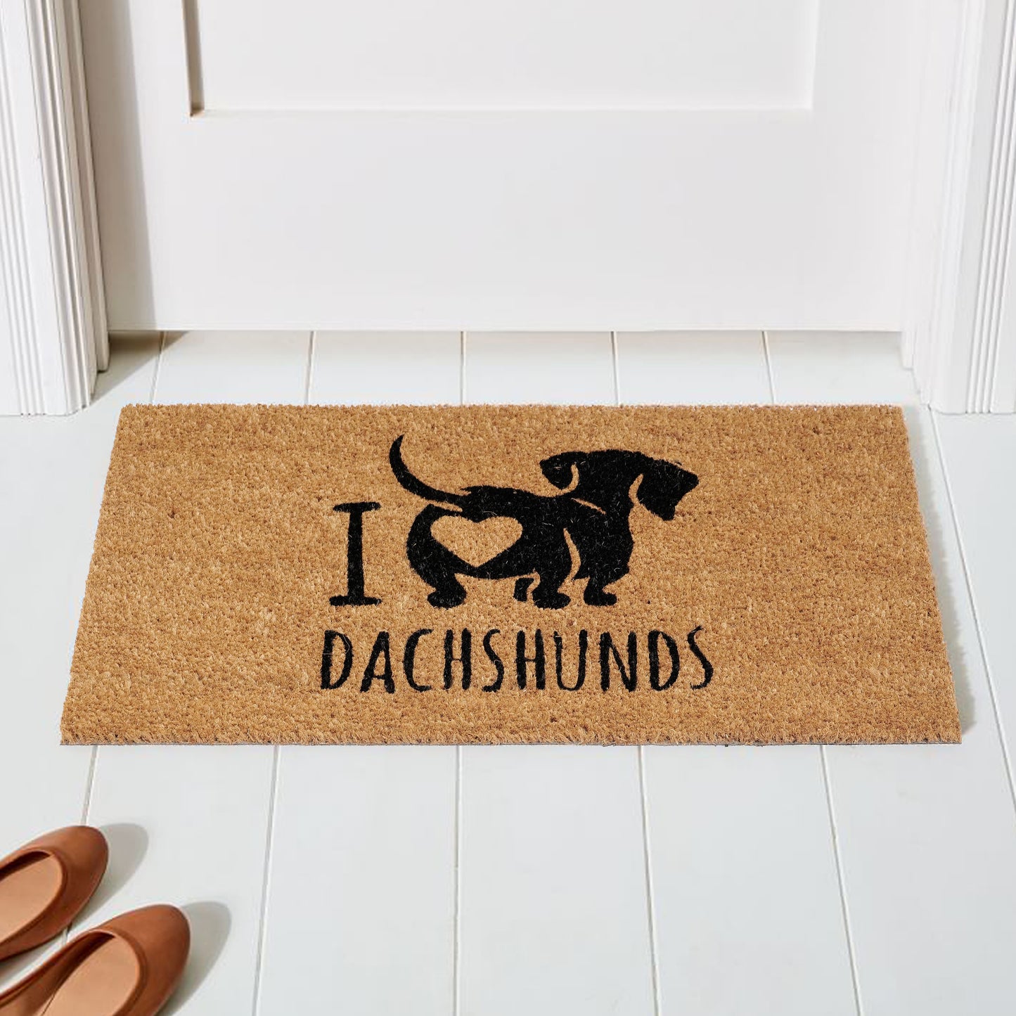 A natural coir doormat with a humorous design of a Dachshund integrated into the text 'I love Dachshunds', placed on a white floor with a pair of shoes at the bottom of the image.