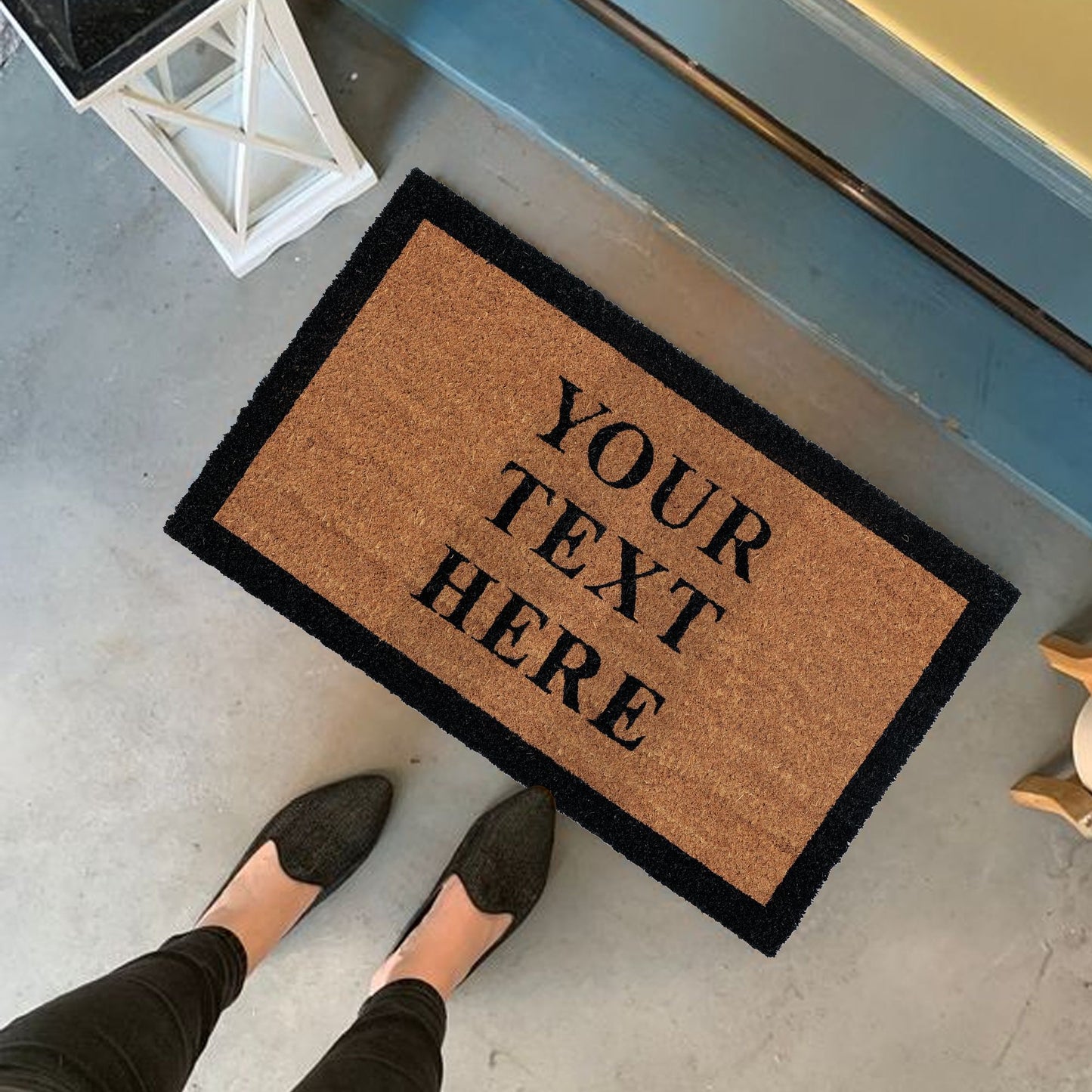 A personalized coir doormat with custom text printed in black. The text 'YOUR TEXT HERE' is displayed on the mat, which is placed on the floor and has a person's feet in black shoes positioned on top of it.