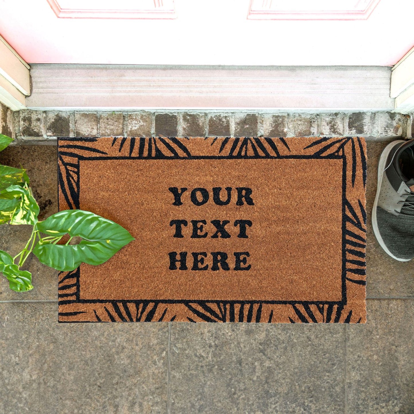 A personalised coir doormat with a tropical leaf border design and custom text 'YOUR TEXT HERE' printed on it. The doormat is placed on the ground with a pair of shoes at the top and a potted plant to the left.