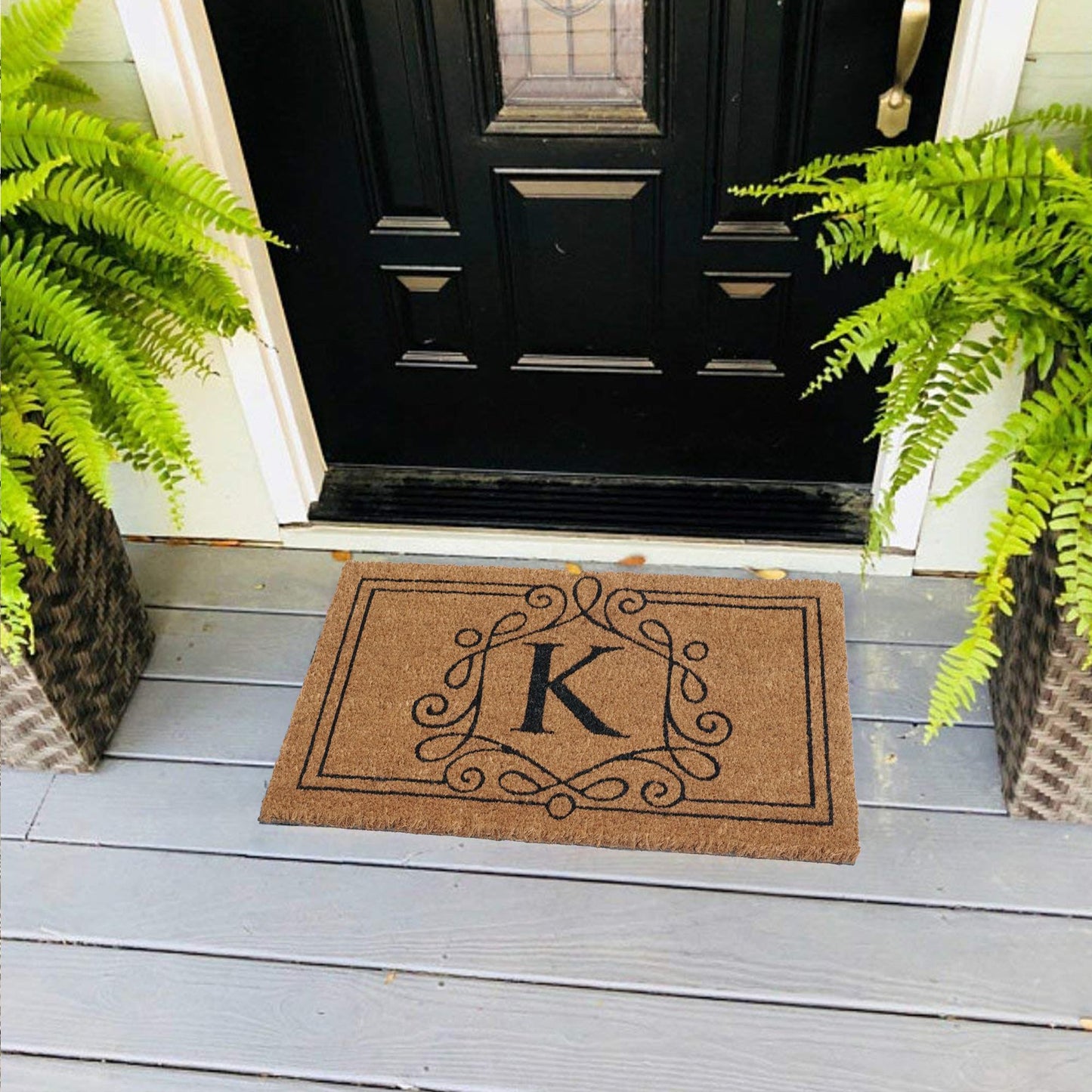 A custom-initial coir doormat with a swirl border design, featuring the initial 'K', placed on a doorstep with green plants on either side.