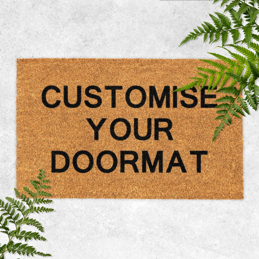 A beige coir doormat with customized black printed text that reads 'CUSTOMISE YOUR DOORMAT', presented on a white background with green foliage around it.