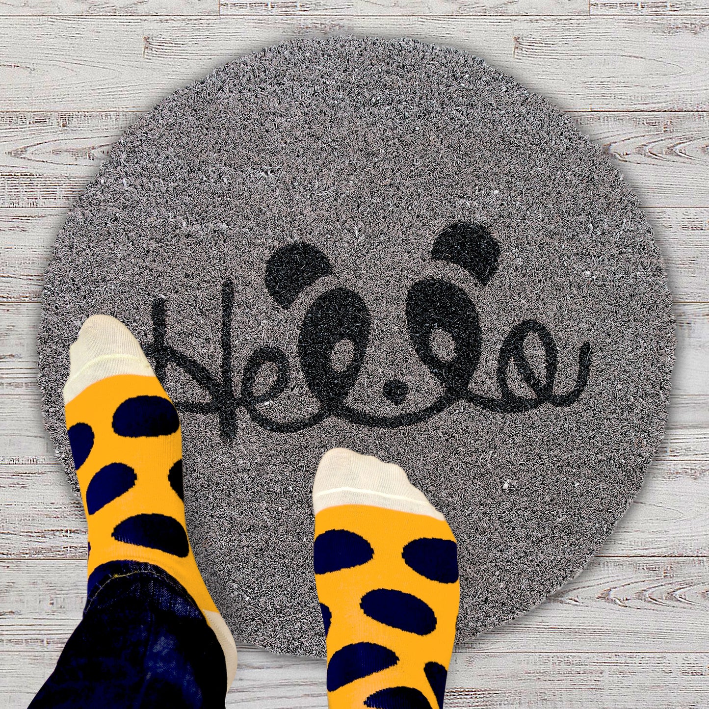 A round-shaped doormat with a grey background and a black and orange design featuring the text 'HELLO' and a panda graphic. A person's feet are visible wearing socks with a similar colour scheme.