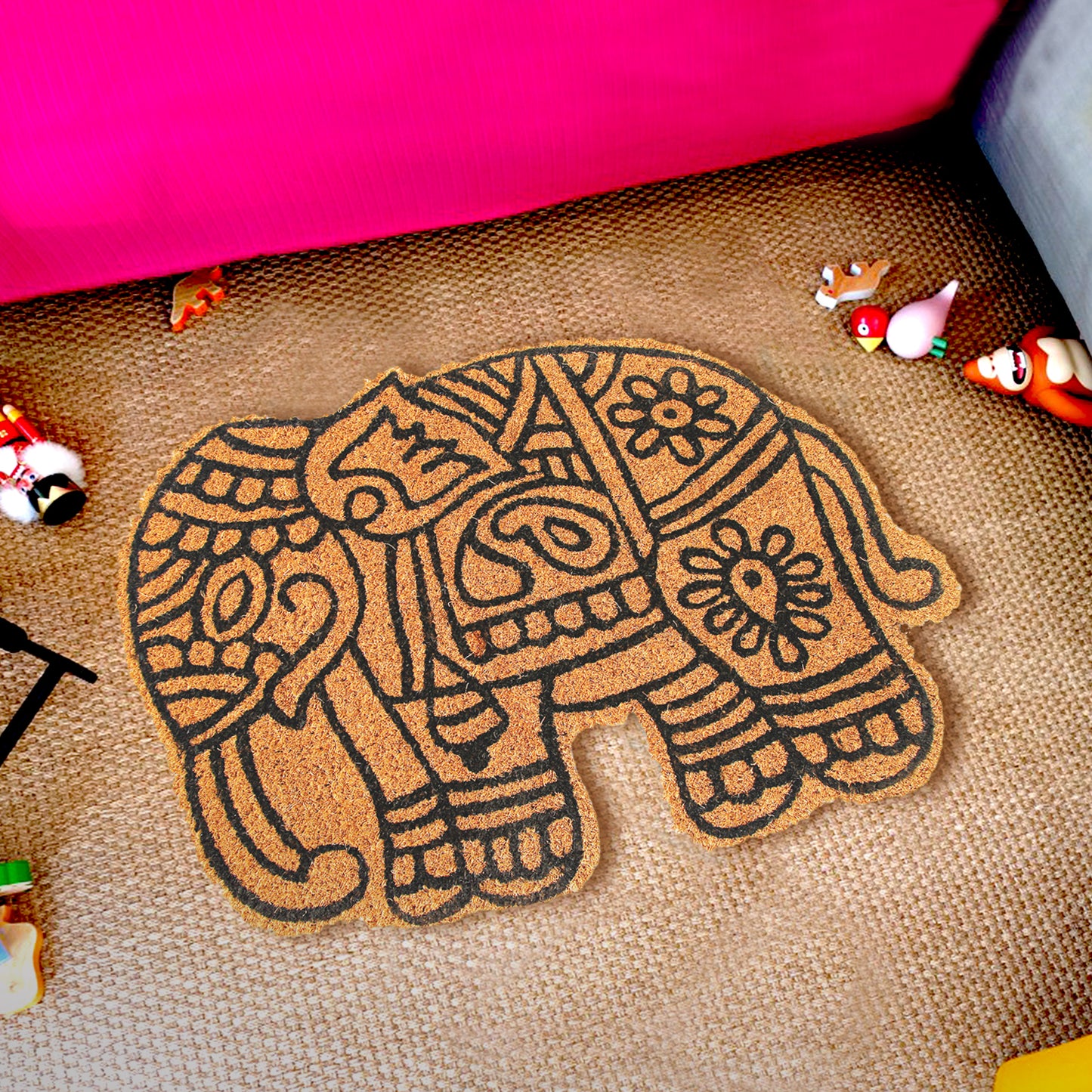 An elephant-shaped coir floor mat with a geometric and floral pattern, placed on a living room floor next to a pink couch with decorative items around it.
