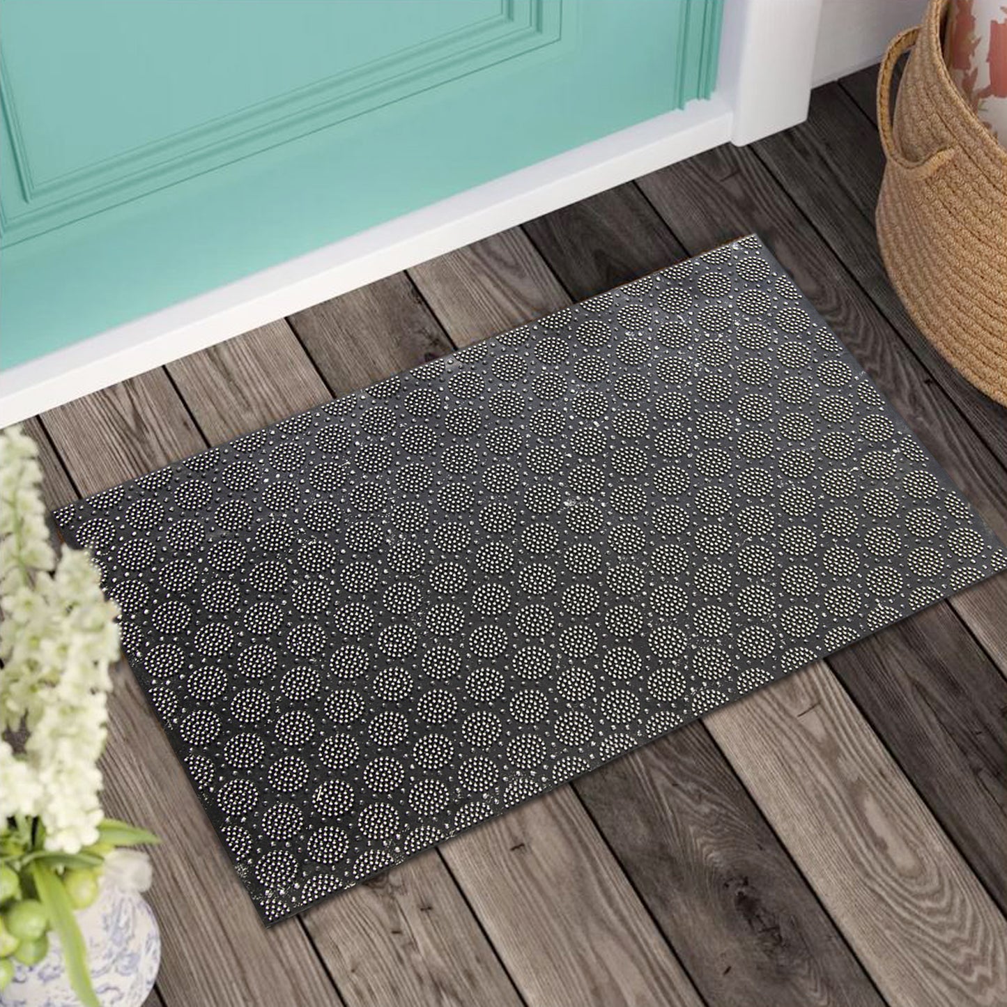 A black rubber door mat with a round geometric pattern, placed on a wooden floor entrance, with a wicker basket and decorative flowers to the side.