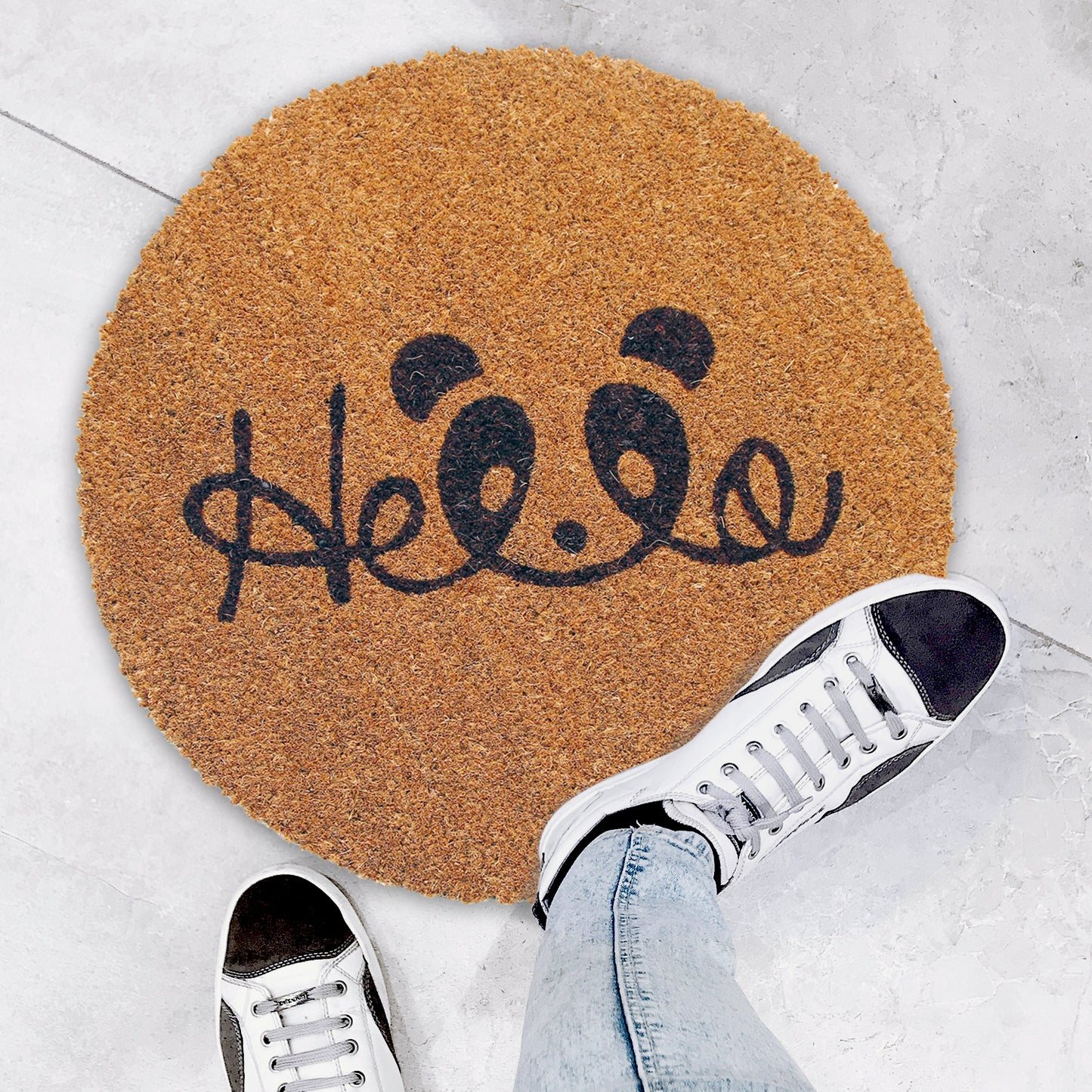A round-shaped natural coir doormat with a black 'Hello' printed design, placed on the floor with a pair of sneakers at its top, and a person's leg and jeans visible to the side.
