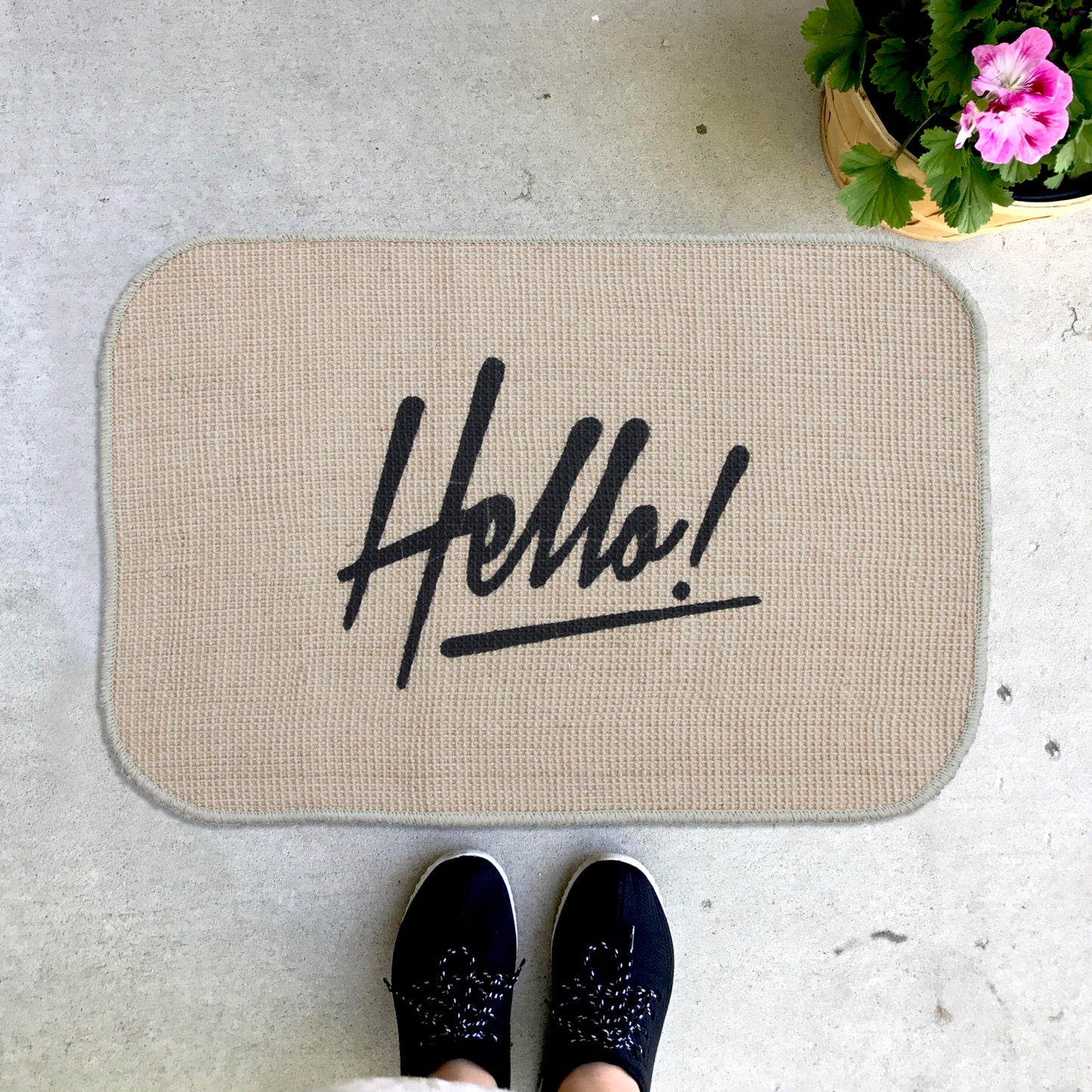 A rectangular beige doormat with the word 'Hello!' printed in black, placed on the floor with a pair of black shoes at the bottom of the image.