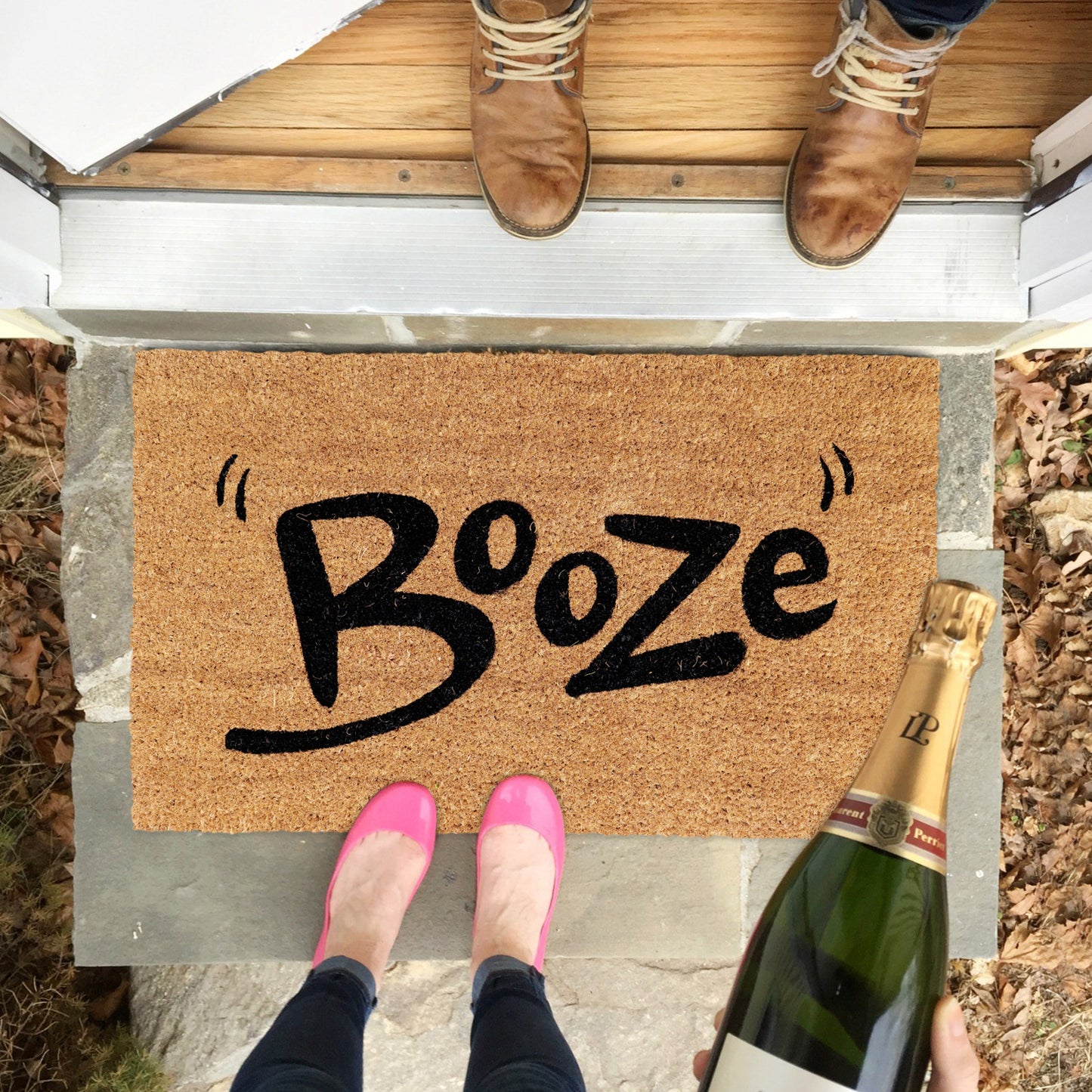 A rectangular natural coir door mat with the word 'BOOZE' printed in black. The mat is placed on the ground, and there are two people standing on it wearing pink shoes.