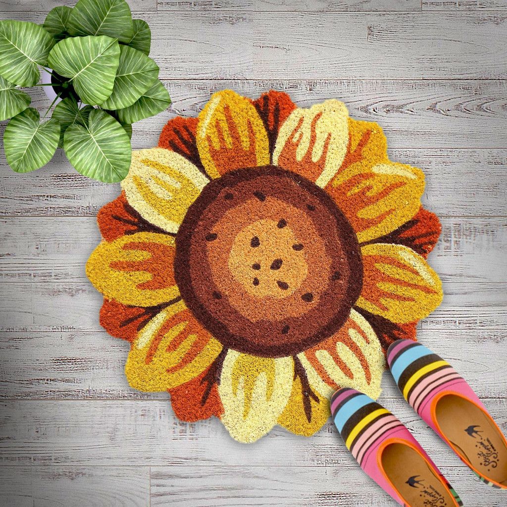 A round natural coir doormat with a sunflower shape and PVC backing, placed on a wooden floor with colorful shoes at the bottom of the image.