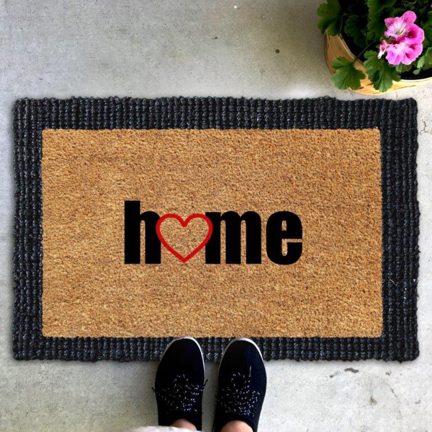 A jute and coir doormat with the word 'Home' printed on it, placed on the floor with a person's feet in shoes on top of it, indicating the mat is for cleaning and welcoming guests.