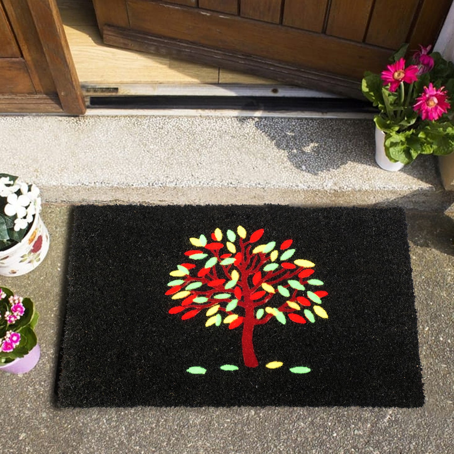A rectangular natural coir doormat with a tree design, placed on the ground in front of a door, with a wooden door frame and potted plants on either side.