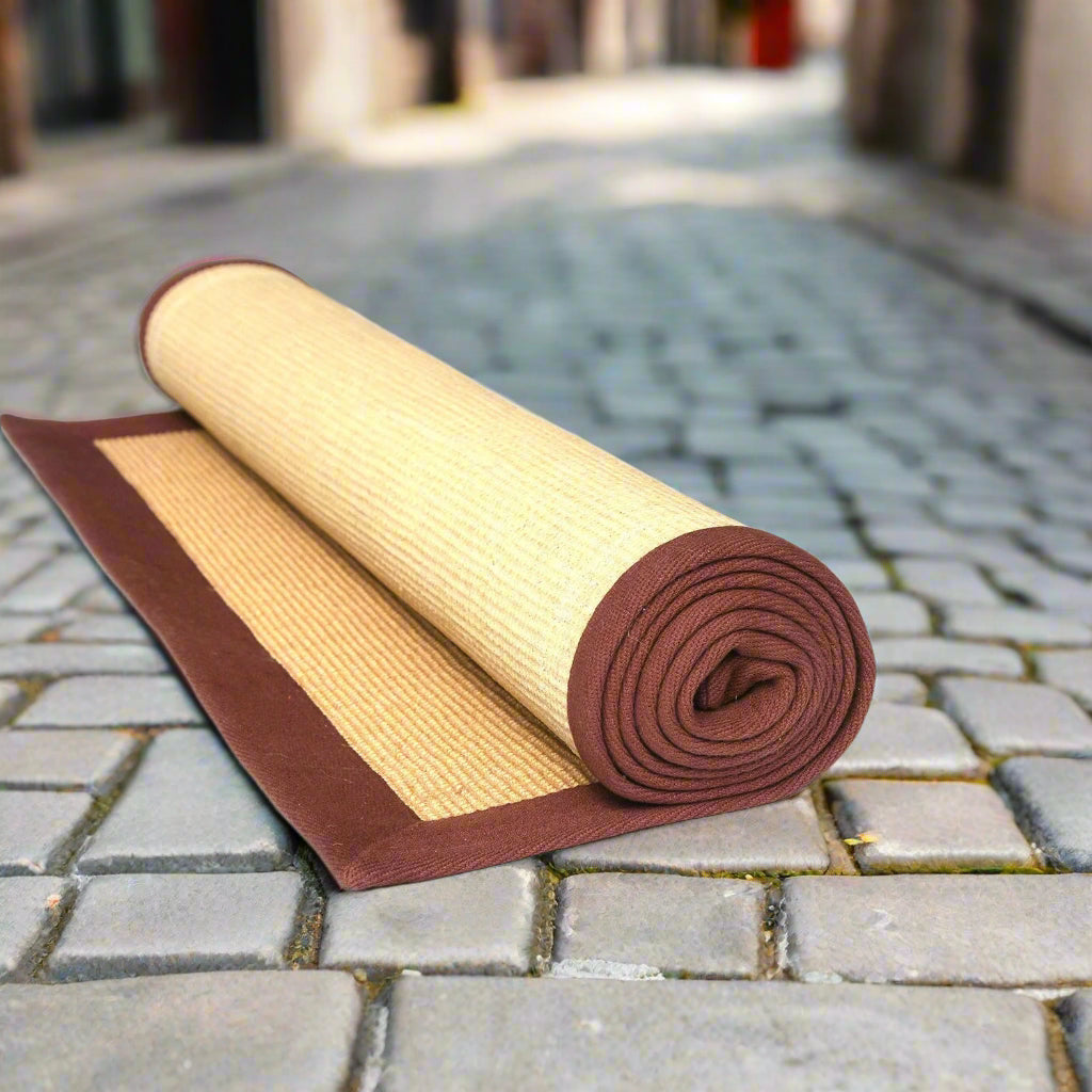 A rolled-up beige yoga mat with a brown cotton border, placed on a cobbled street.