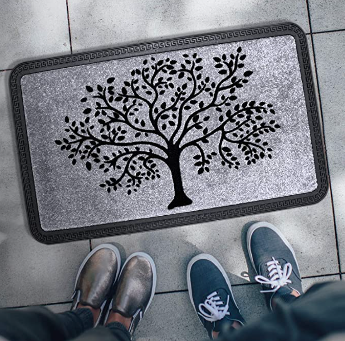 A rectangular doormat with a black floral tree design on a grey background, placed on the floor with a pair of shoes at the top of the image.
