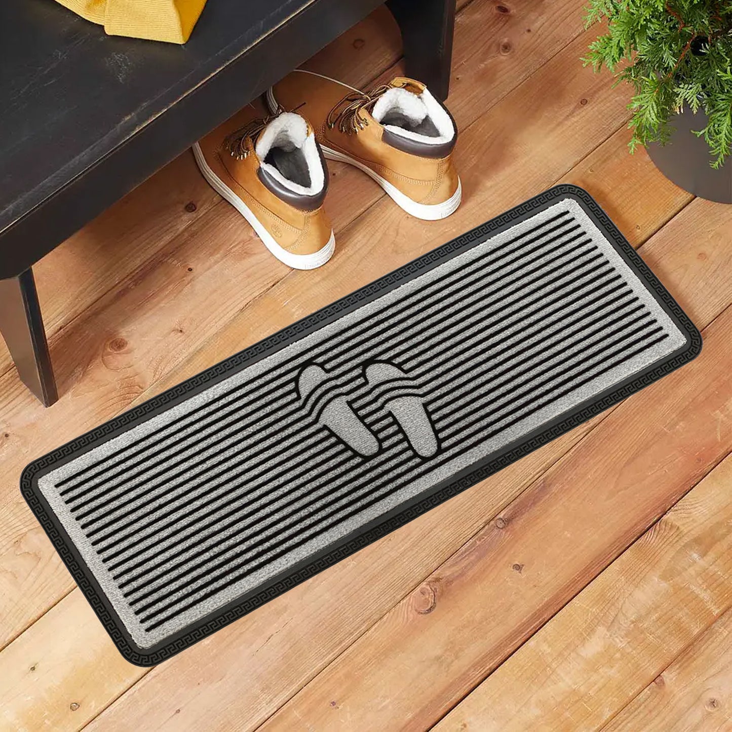 A rectangular, grey polypropylene doormat with a flocked design featuring a pair of feet, placed on a wooden floor. There are shoes atop the mat, and a potted plant and a black bench are visible in the background.