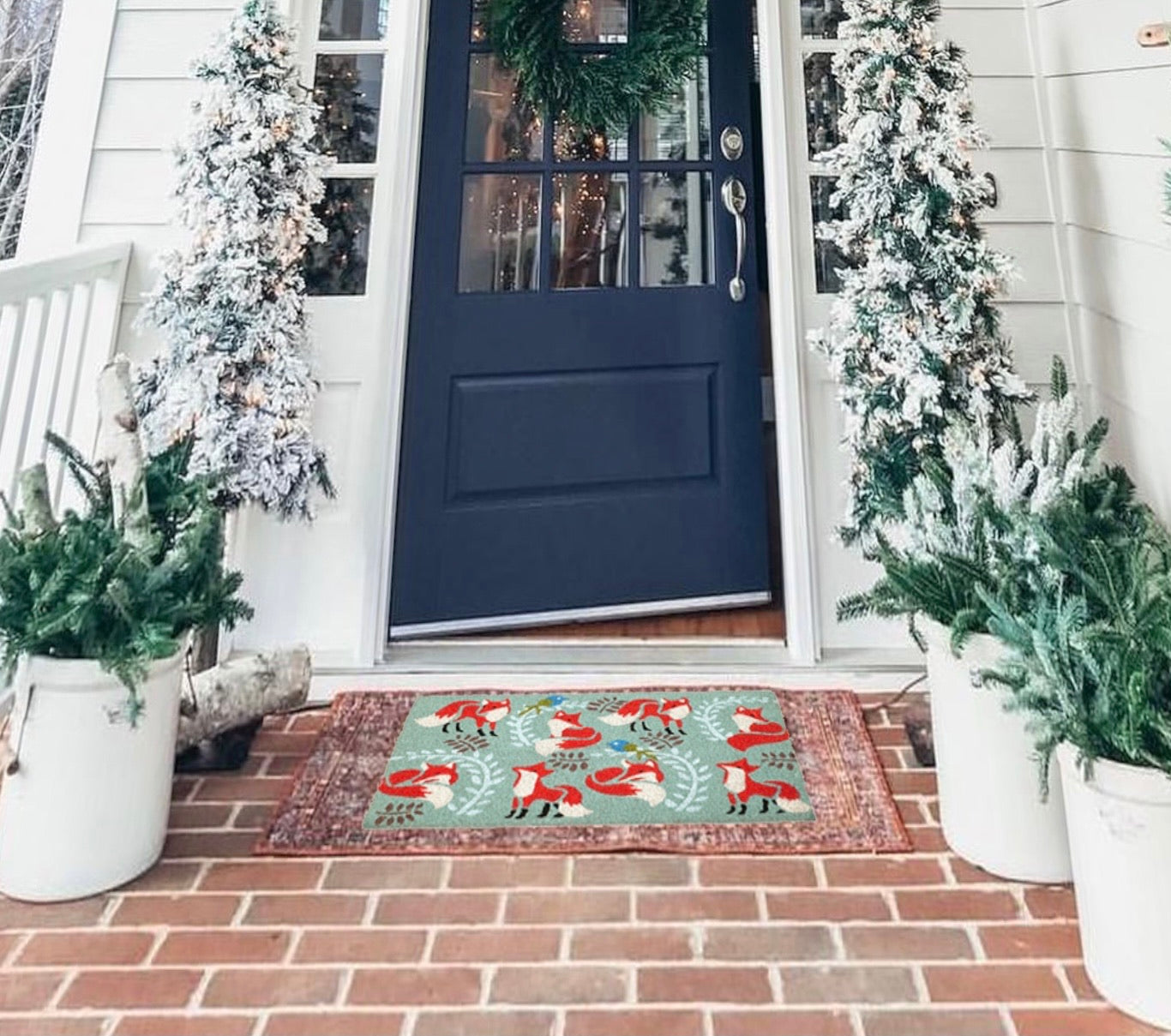 A natural coir doormat with a Christmas-themed pattern, featuring a fox design, is placed on the doorstep of a home with a decorative border and potted plants on either side.