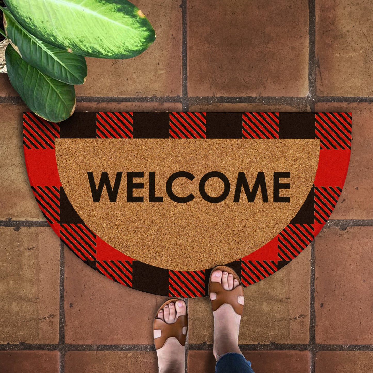 A natural coir doormat with a black and red checkered pattern and the word 'Welcome' printed on it, placed on a tiled floor with a person's feet in sandals stepping on it.