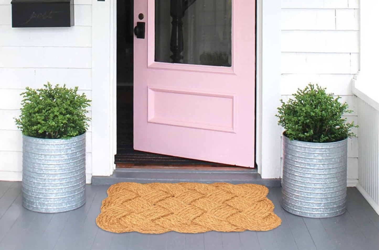 Pink front door with two potted plants and a textured doormat on a gray porch.