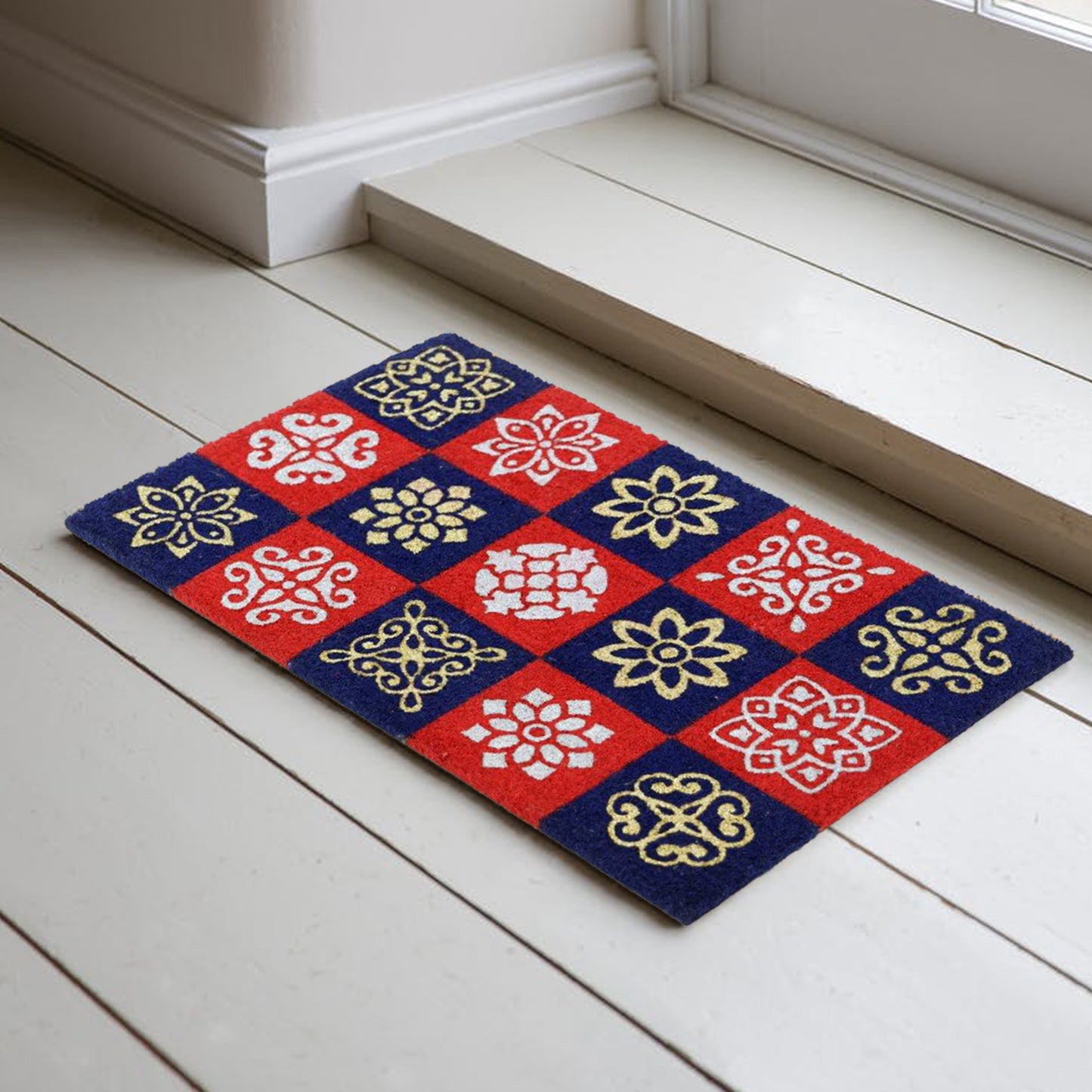 A rectangular natural coir door mat with a vibrant red and blue tile design and floral patterns, placed on a white floor by a door.