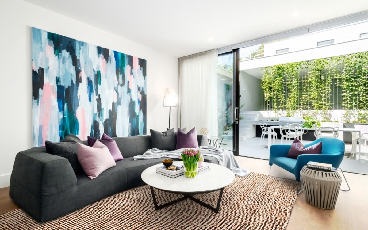 A modern living room featuring a grey sofa with purple cushions, a white round side table with a lamp, a woven jute rug, and a large abstract painting on the wall.