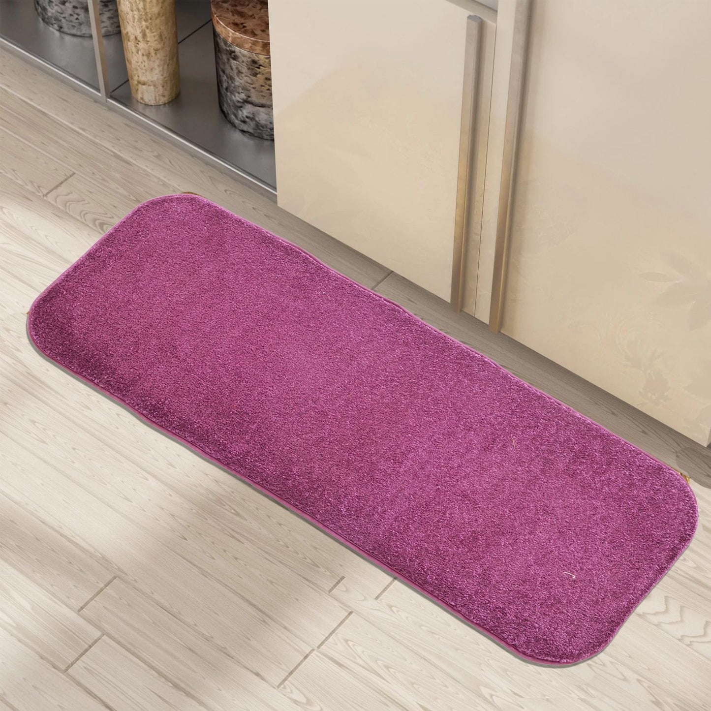 A rectangular purple doormat with a textured surface is placed on a light floor, with a kitchen cabinet and shelves in the background.