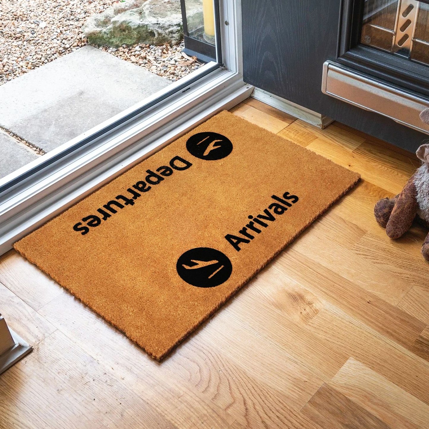 A rectangular, natural coir entrance doormat with black flocked design and the text 'ARRIVALS' printed on it, placed on a wooden floor by a door.