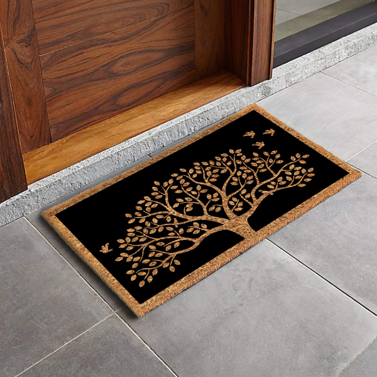 A rectangular natural coir black door mat with a printed Tree of Life and chirping birds design, placed on a tiled floor.