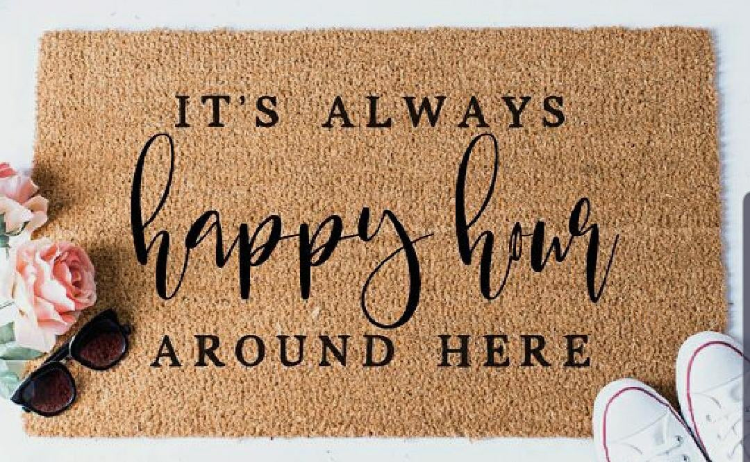 Rectangular natural coir floor mat with a printed design that reads 'It's always happy hour around here' in cursive script, placed on a white surface with a pair of sunglasses and a shoe on top of it.