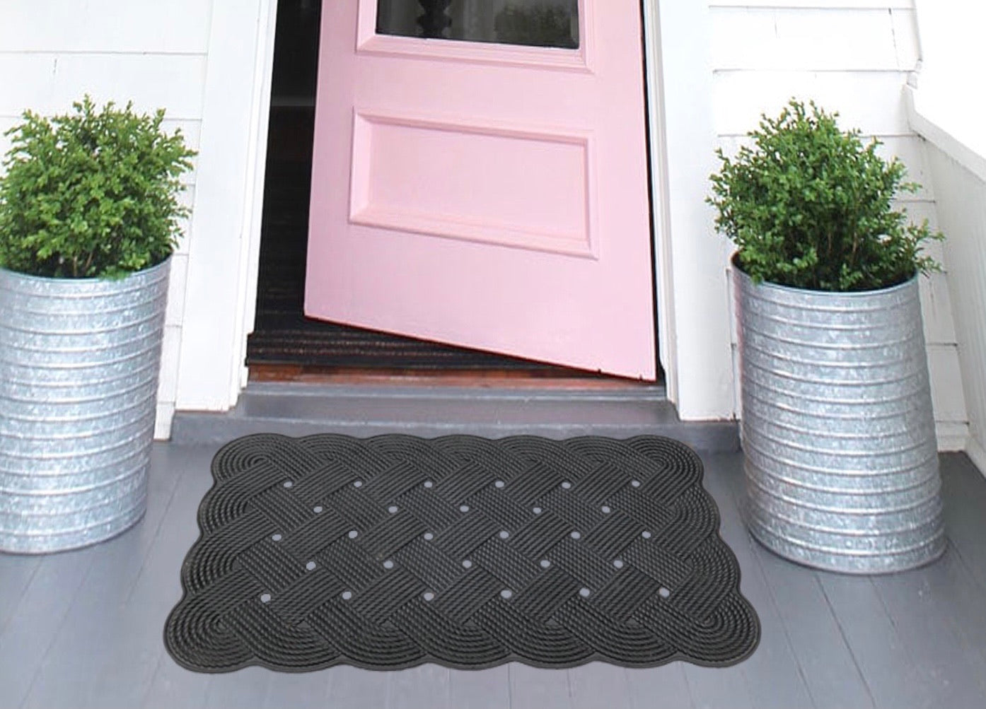 Rubber mats with a knot design pattern, in black colour, placed on a doorstep with a pink door and green shrubbery in the background.