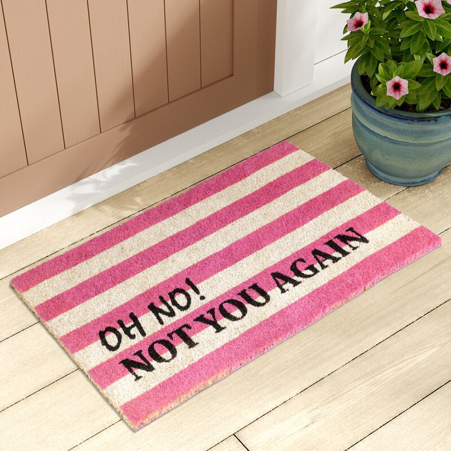 A rectangular doormat with pink and white retro stripes and the text 'OH NO NOT YOU AGAIN' printed on it, placed on a wooden floor by a door.
