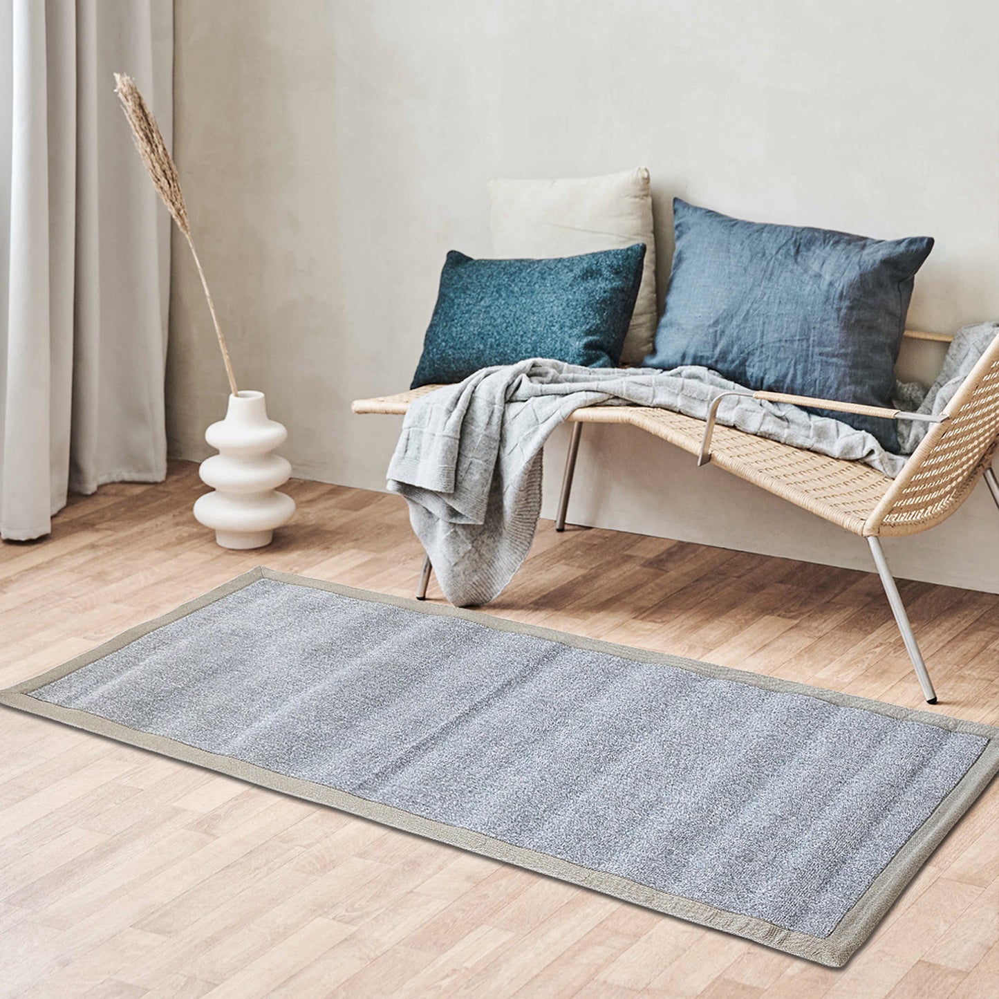 A grey yoga mat with a cotton border laid out on the floor in a living room setting, next to a modern chair and a window with light coming through.