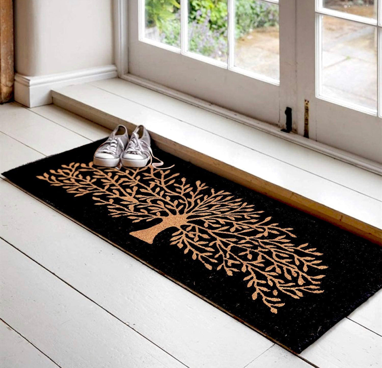 A black coir floor mat with a brown long tree design, placed on a white floor with a pair of shoes on top of it, near a window.
