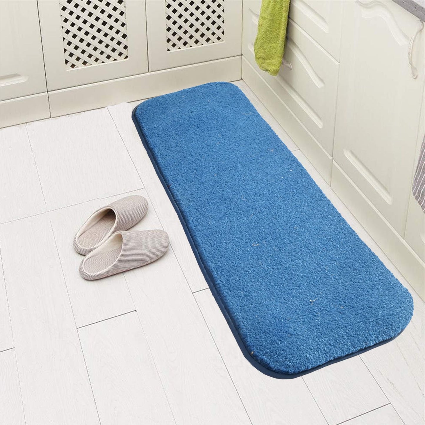 A blue rectangular doormat with a non-skid surface, placed on a white tiled floor next to a pair of gray slippers.