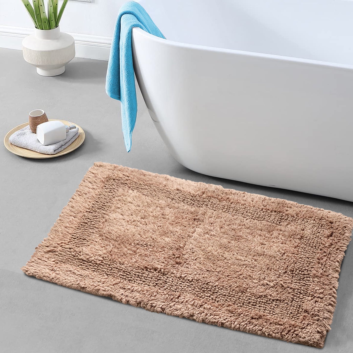 A brown cotton bath mat with a textured surface, placed on a tiled floor next to a bathtub with a blue towel and a wooden holder with a white candle.