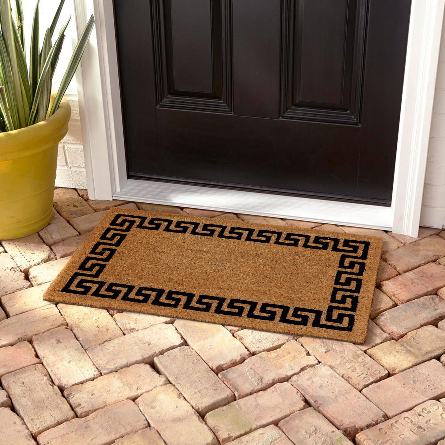 A rectangular natural coir floor mat with a geometric Greek pattern border printed on it, placed on a brick pathway leading up to a door.