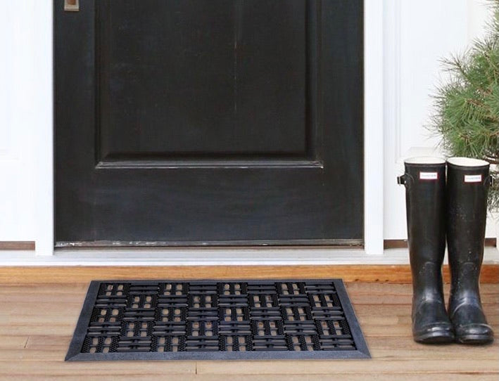 A black criss-cross rubber pin door mat is placed on a wooden floor in front of a door, with a pair of boots at the top of the mat and a pine tree in the background.