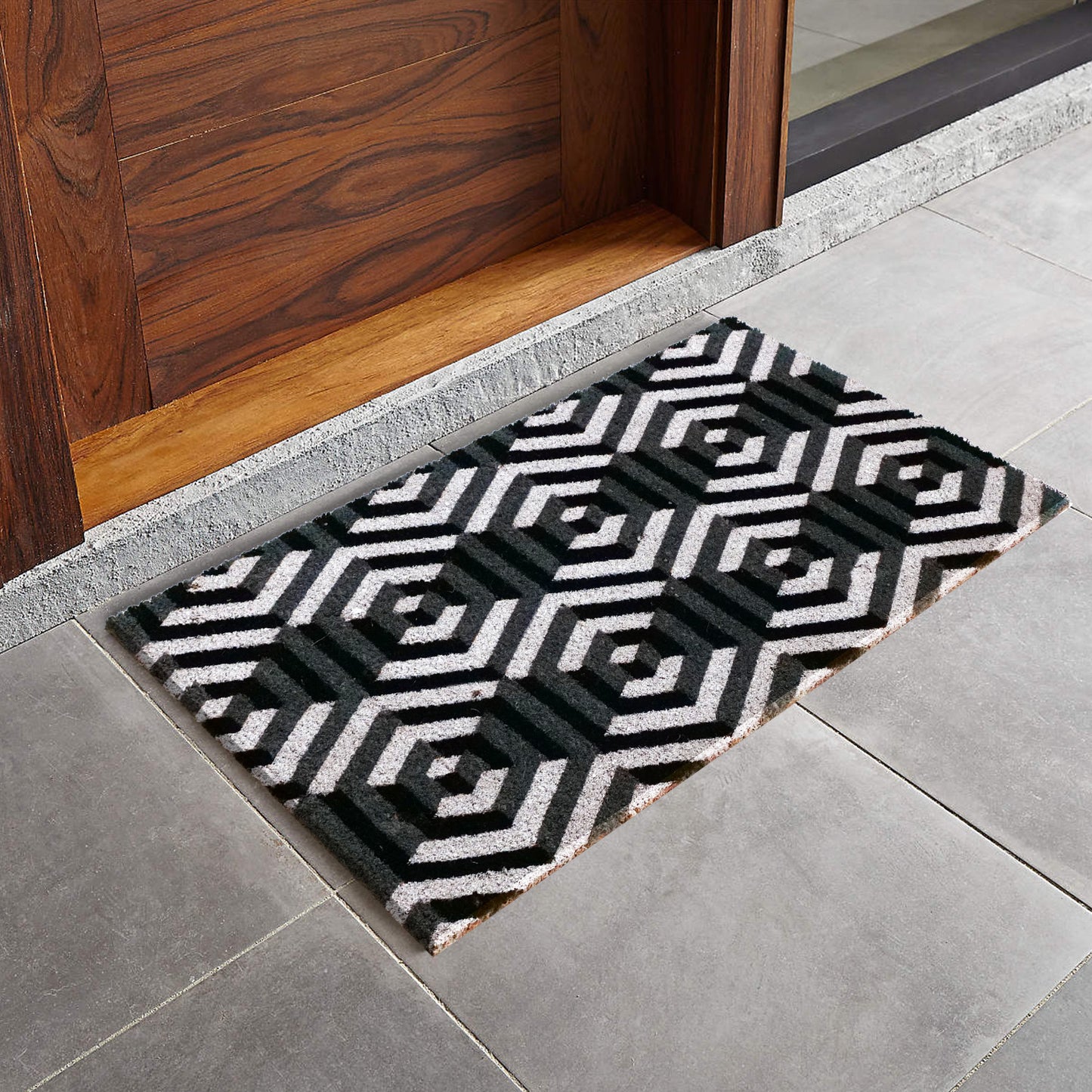 A stylish natural coir doormat with a geometric 3D octagon design, placed on a tiled floor in front of a wooden door, featuring a black and white colour scheme.