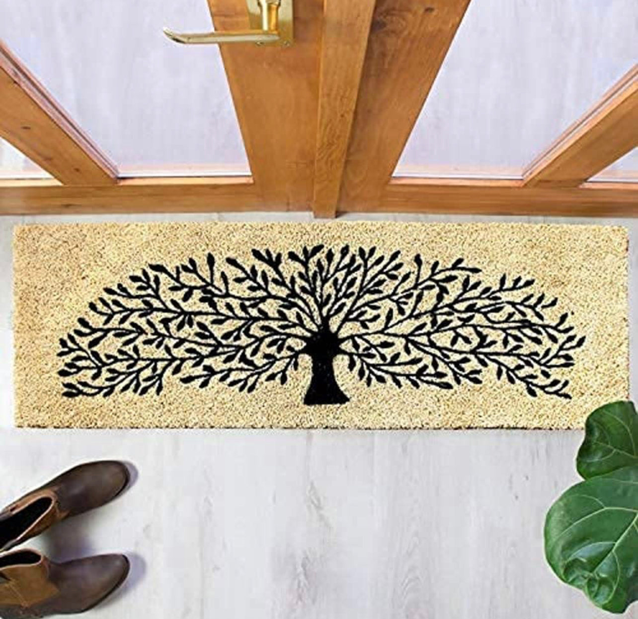 A beige and black tree design coir doormat placed on the floor, with a pair of boots at the bottom of the image and a plant to the side.