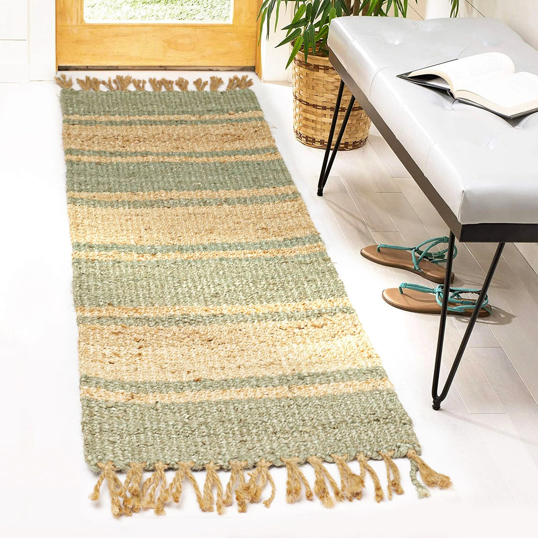 A flatweave jute rug with fringes in natural and pastel green shades, placed on a white floor in a home environment, near a door, with a bench and a plant beside it.