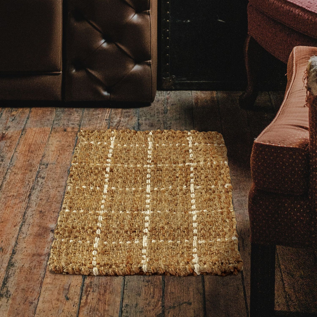 A natural beige jute rug with a plaid pattern, placed on a wooden floor in a room with a brown leather sofa and a floral-patterned armchair.