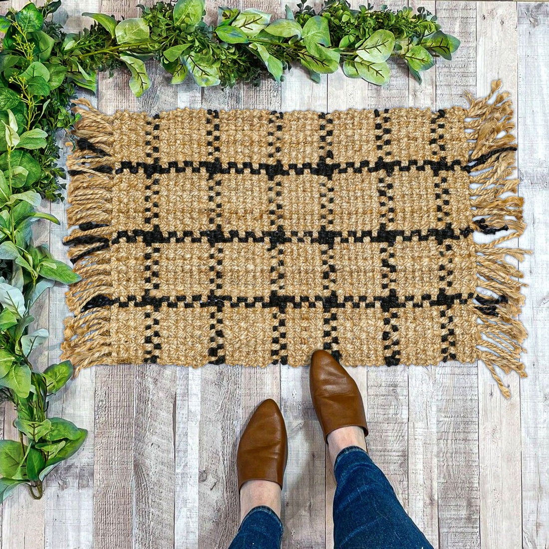A rectangular jute rug with a plaid pattern in beige and brown tones, featuring a shaggy texture, is placed on a wooden floor with green plants in the background and a pair of brown shoes at the bottom of the image.