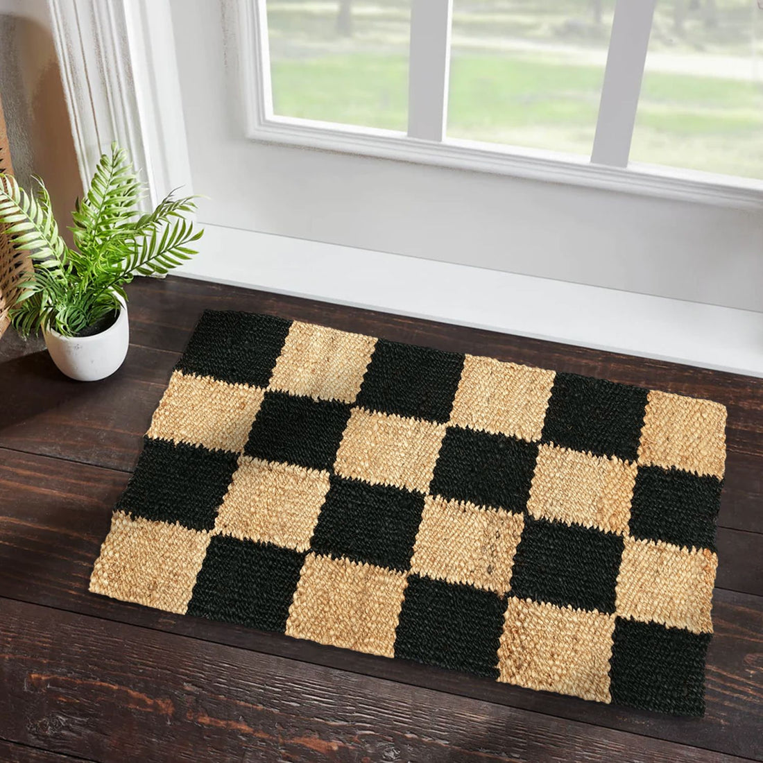 A rectangular jute rug with a black and beige checkered pattern, placed on a wooden floor next to a window with a view of greenery.