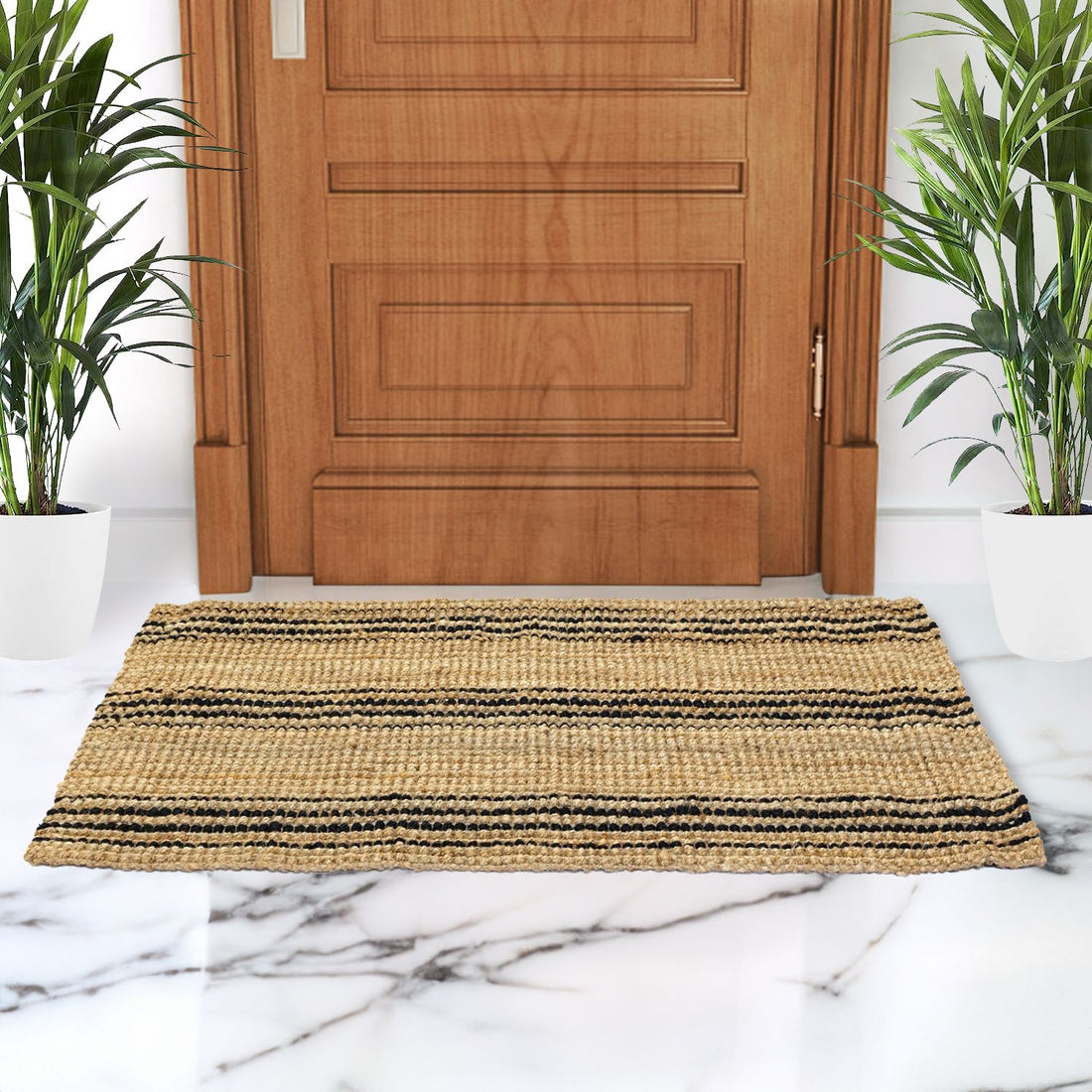 A handwoven jute rug with a geometric pattern, primarily in beige and brown tones, is placed on a marble floor with a wooden door in the background and two potted plants on either side.