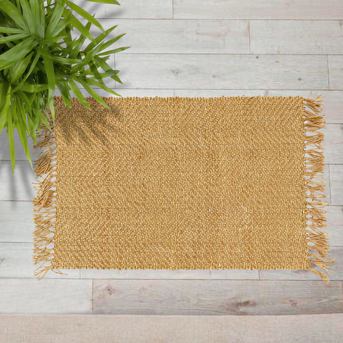 A rectangular jute rug with a fringed edge, placed on a wooden floor, with a green plant in the background.