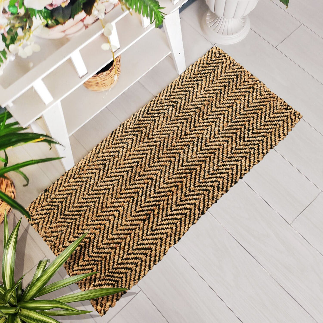 A rectangular jute rug with a herringbone weave pattern, placed on a light grey floor, with a white chair and a potted plant beside it.