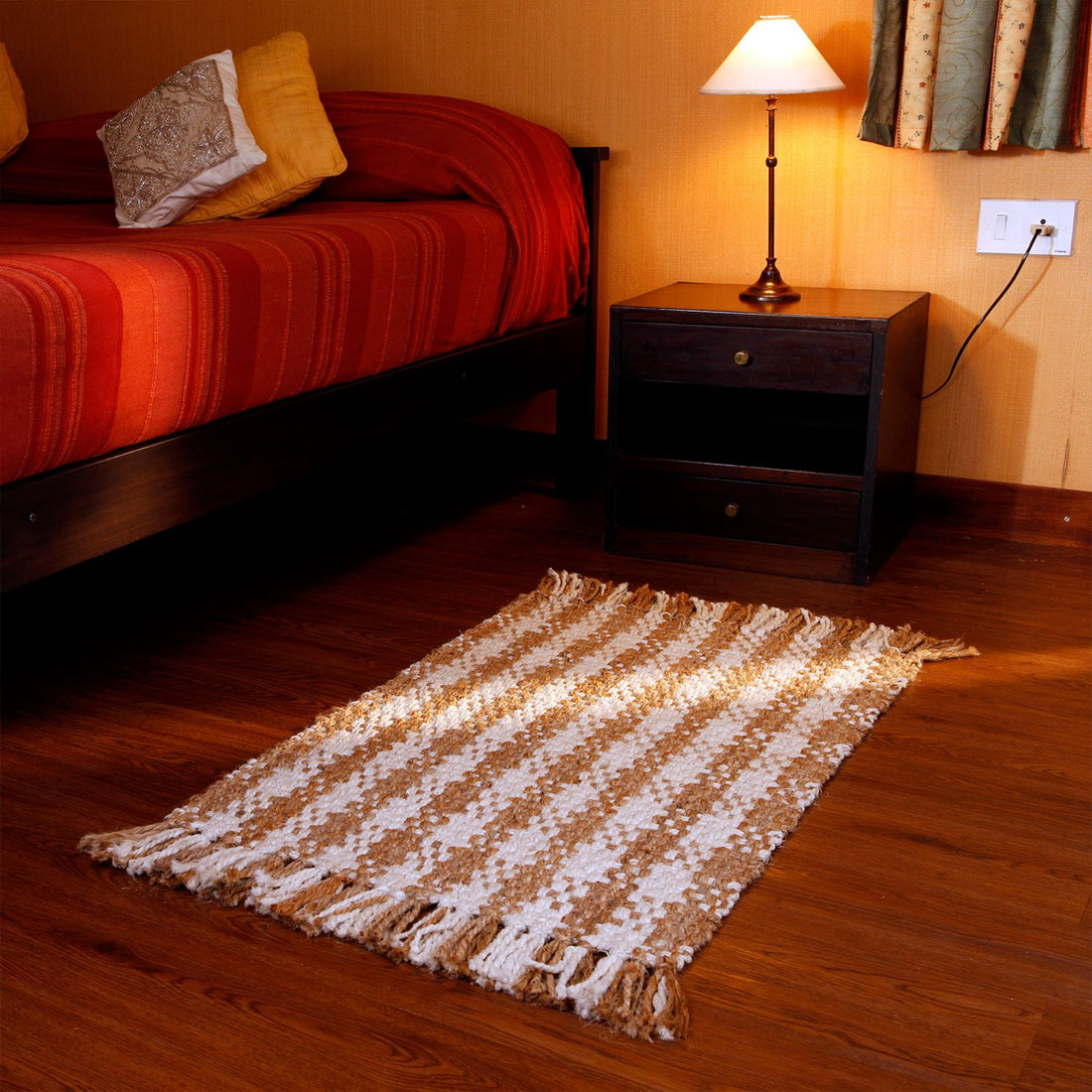 A handmade jute carpet with a geometric pattern, in a neutral colour, is placed on a wooden floor next to a bed with orange bedding and a nightstand with a lamp.