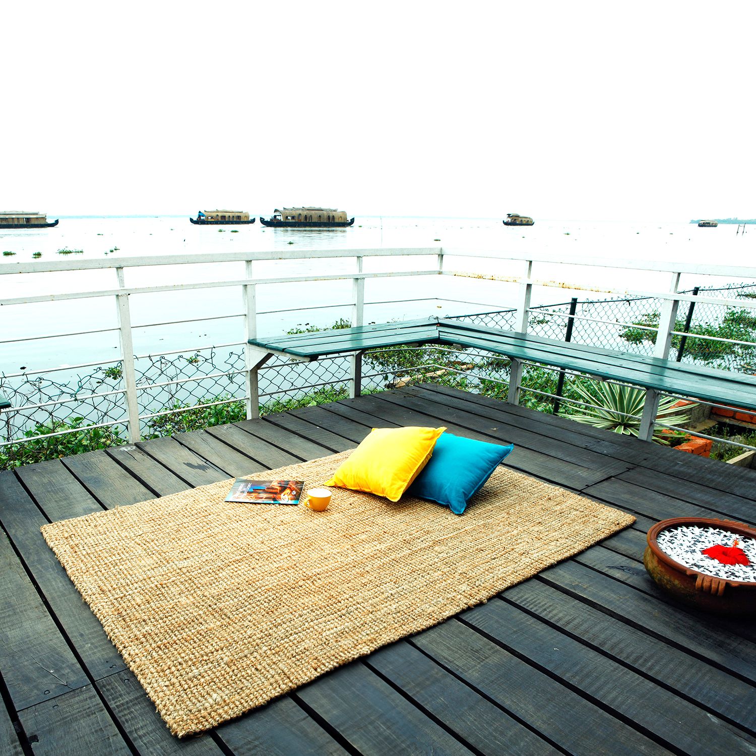 A handmade jute rug with a flat-weave pattern is placed on a wooden deck with a scenic view of a body of water and houseboats in the background. There is a pillow on the rug, and a book is placed on top of the pillow.