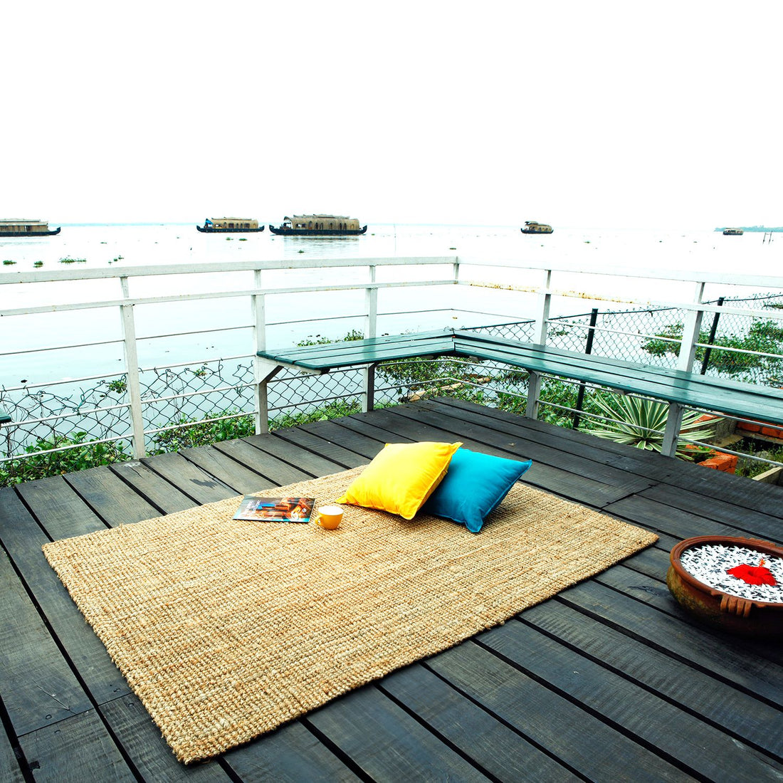 A handmade jute rug with a flat-weave pattern is placed on a wooden deck with a scenic view of a body of water and houseboats in the background. There is a pillow on the rug, and a book is placed on top of the pillow.
