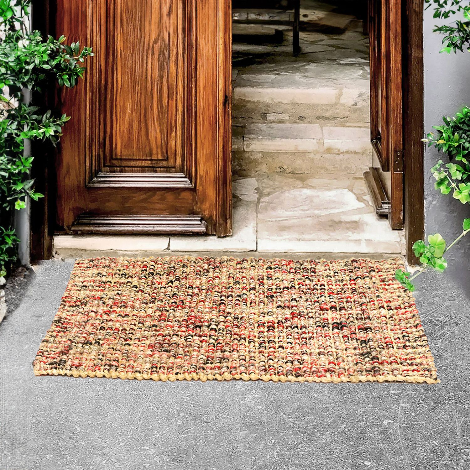 A handwoven jute carpet with a red and black tie-dye pattern, placed on the floor in front of a wooden door, with plants and stairs in the background.